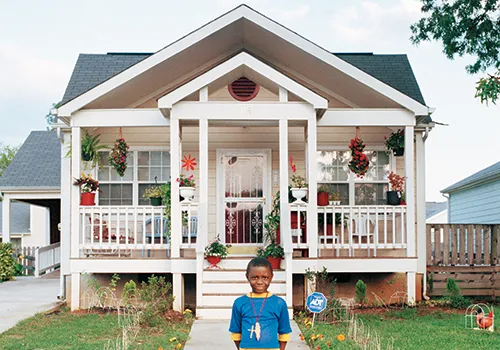 An all-American styled home front yard and porch view with a young African American boy standing in front, wearing a blue shirt.