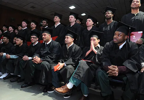 Young men shown sitting together, wearing graduation caps and gowns, in anticipation for their graduation ceremony.