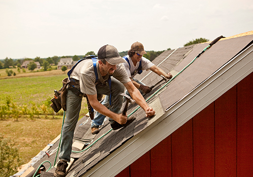 Two construction workers up on a barn rooftop, shingling tiles together.