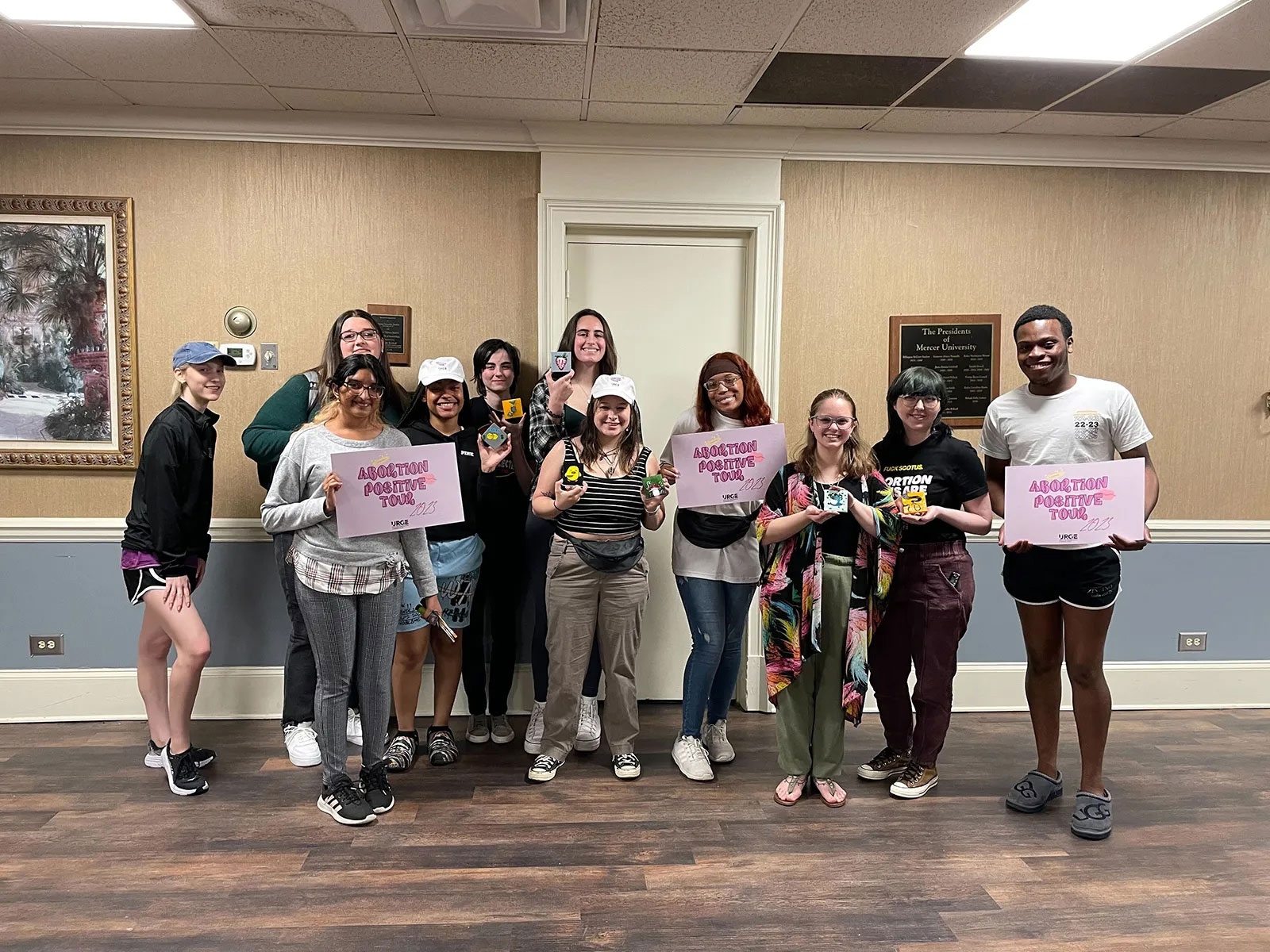 A group of diverse young activists stand together indoors, holding pink signs that read "Abortion Positive Tour 2023." Some individuals also hold small, colorful boxes. This image represents youth advocacy, reproductive rights movements, and campus-based social justice organizing.