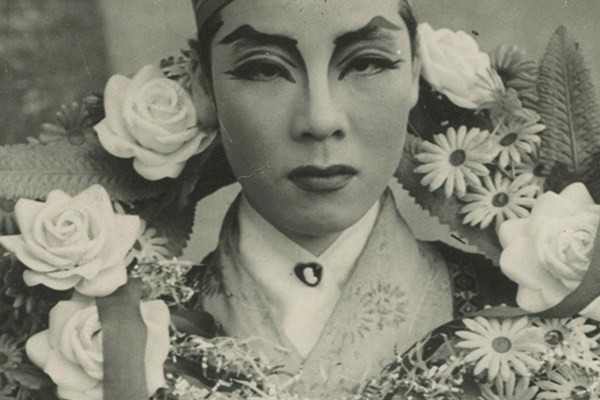 A gray-scale photograph of a Yeoseong Gukgeuk (National Women's Theater) performer gazing forward with prominent makeup. There is a wreath of flowers hanging behind their head and around their chest.