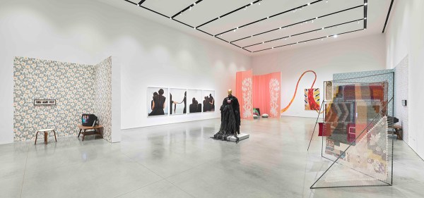 A white gallery interior with cement floors. There are multiple artworks in the gallery including a living room-inspired installation with a vintage television and floral wallpaper; a row of four photographs of women braiding their hair; a gold mannequin in a black dress, a pink curtain installation, a colorful canvas with paint.