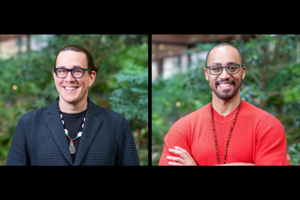 Two people smiling in front of a garden background. Both people are wearing necklaces; one is wearing a black blazer and the other a red sweater.