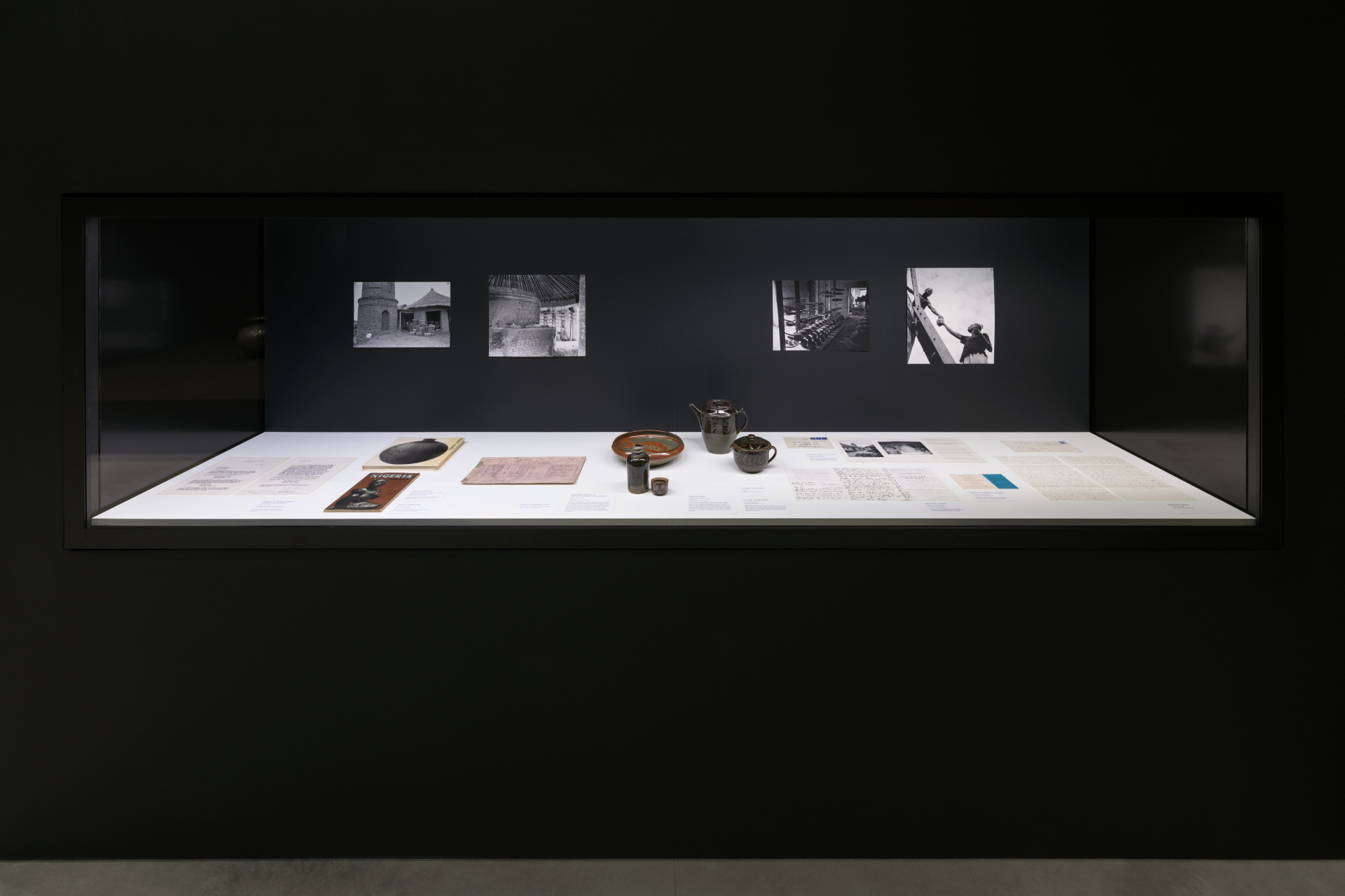 A glass vitrine holding archival paper objects, ceramic works, and historic photography documentation of a pottery center.