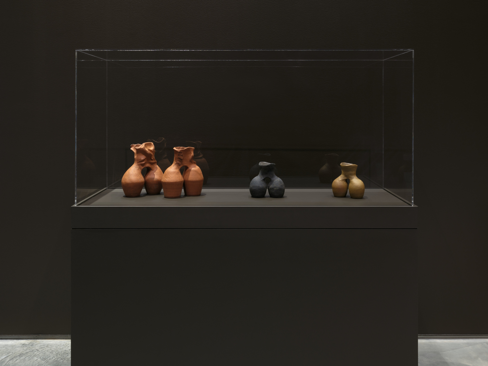 A glass vitrine holding four ceramic sculptures that each have a similar two-barrel shape, but made of different ceramic material and of slightly different heights.