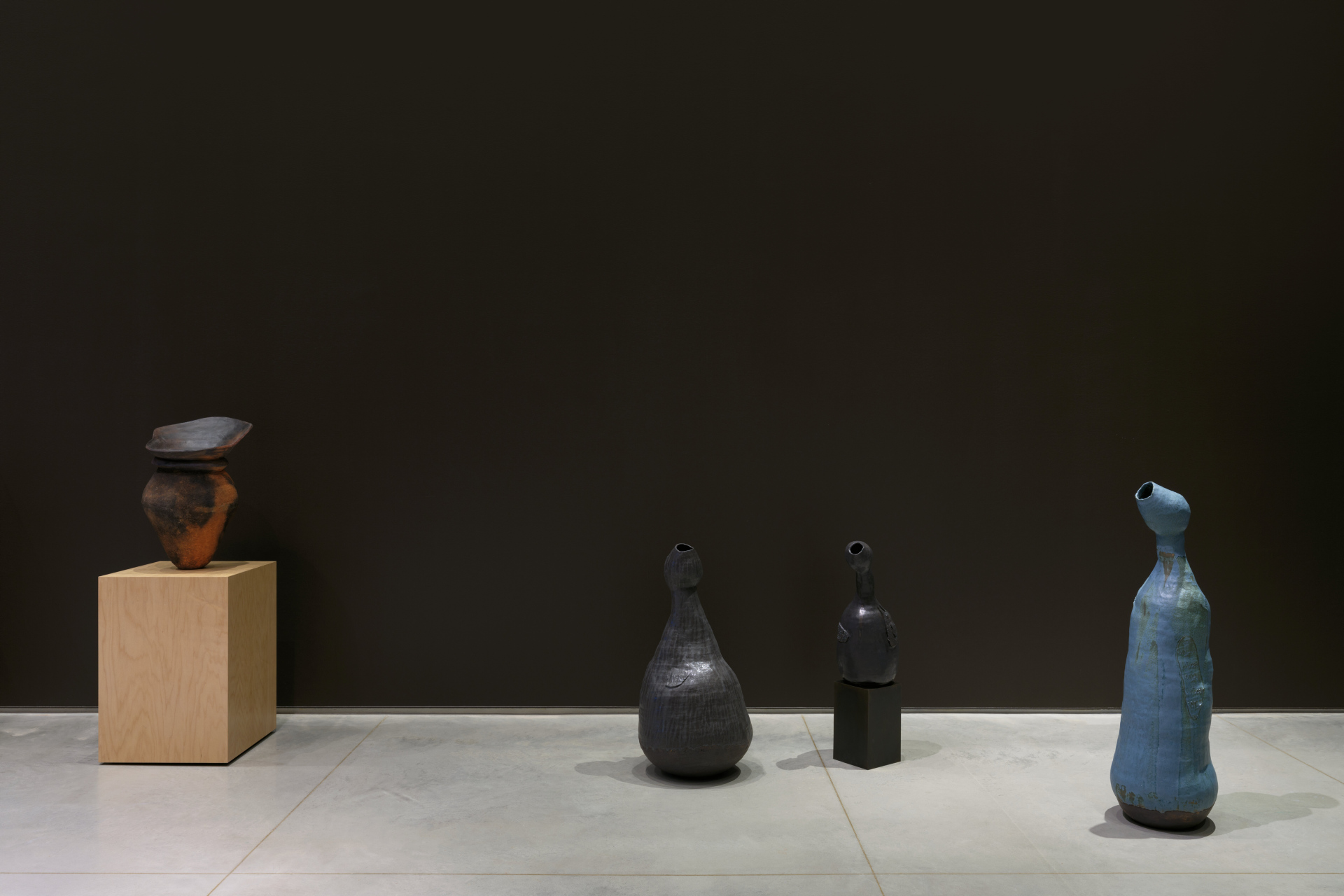 An exhibition showing a ceramic vase on a pedestal, and three clay sculptures with lumpy and irregular bodies.