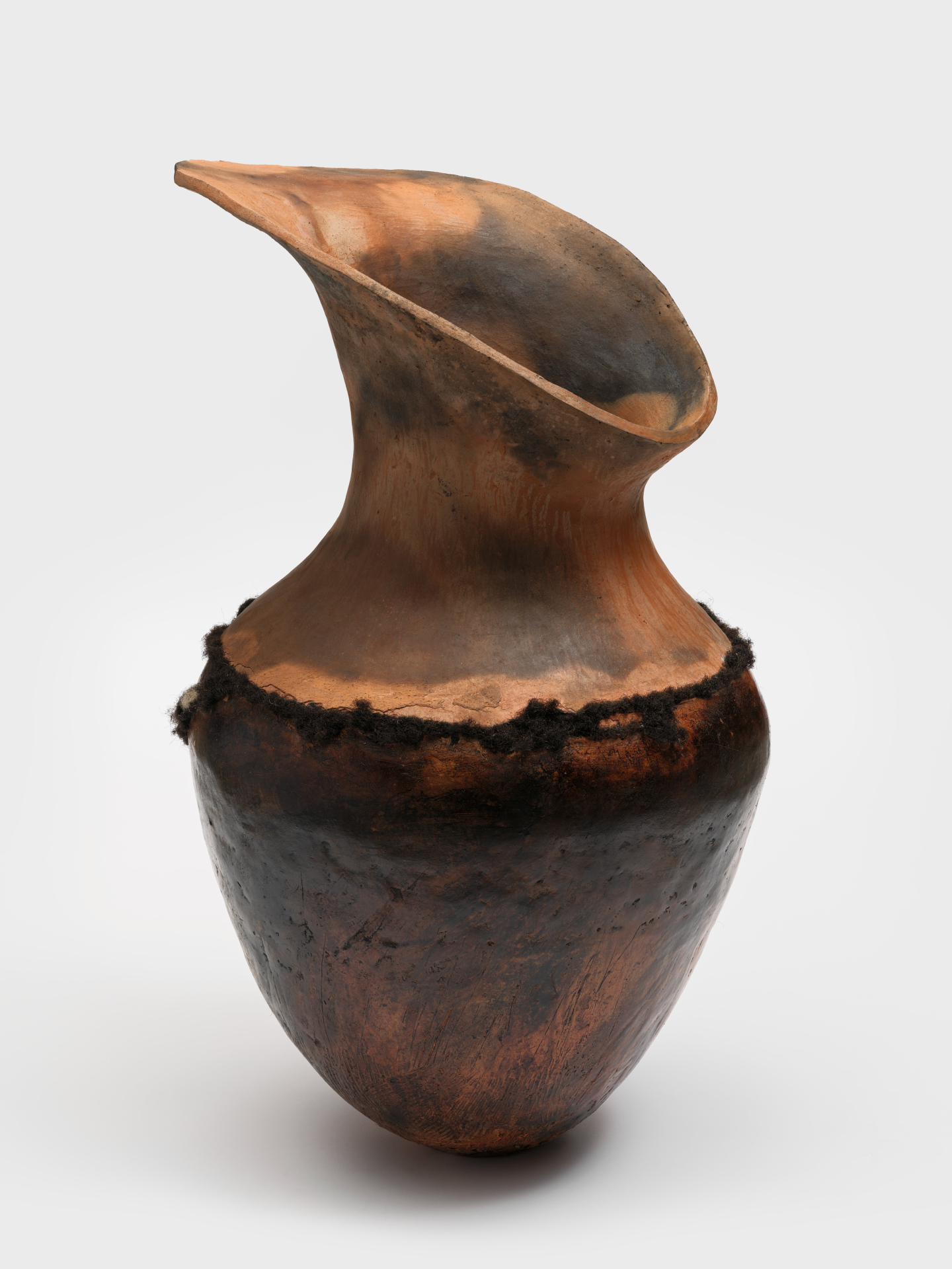A large pit-fired ceramic pot with a wide teardrop-shaped mouth and dark curly hair belted around the vessel.