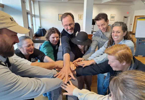 A group of nine smiling individuals place their hands together in a central huddle indoors. This image captures community building, collective action, and local civic engagement, illustrating the "infrastructure of trust" within rural American grassroots democracy movements.